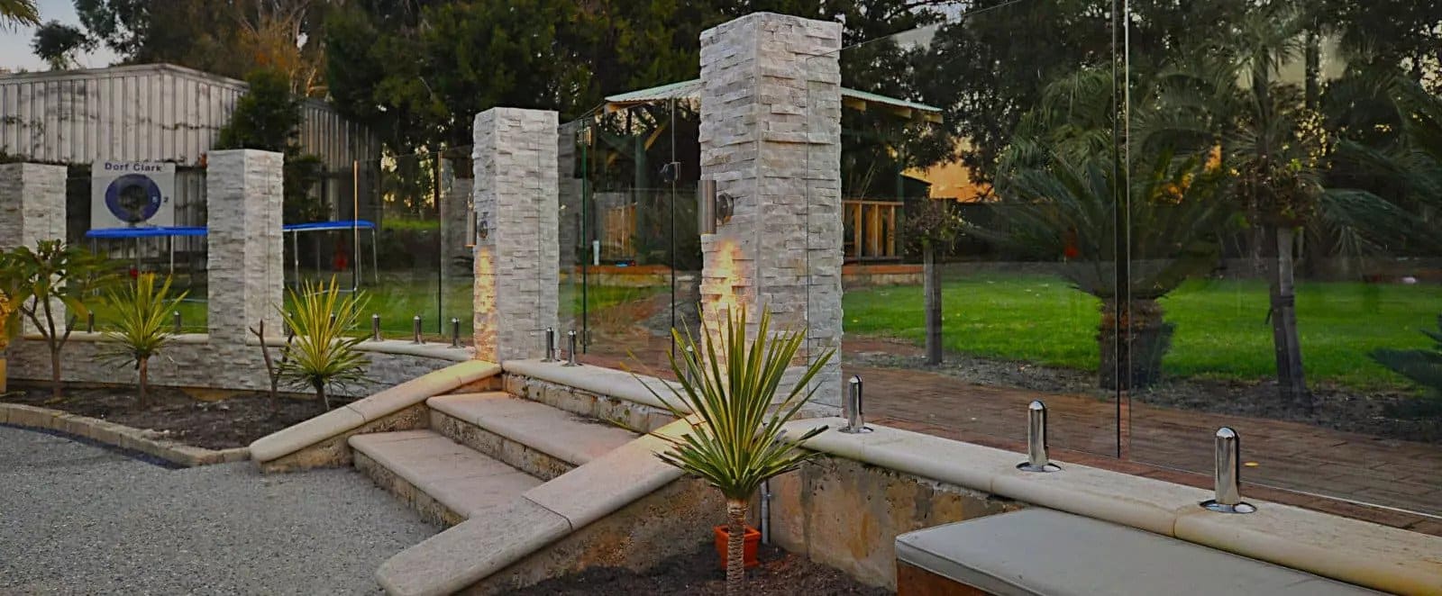 Elegant backyard featuring illuminated stone pillars, glass fencing, and landscaped greenery for a serene outdoor space.