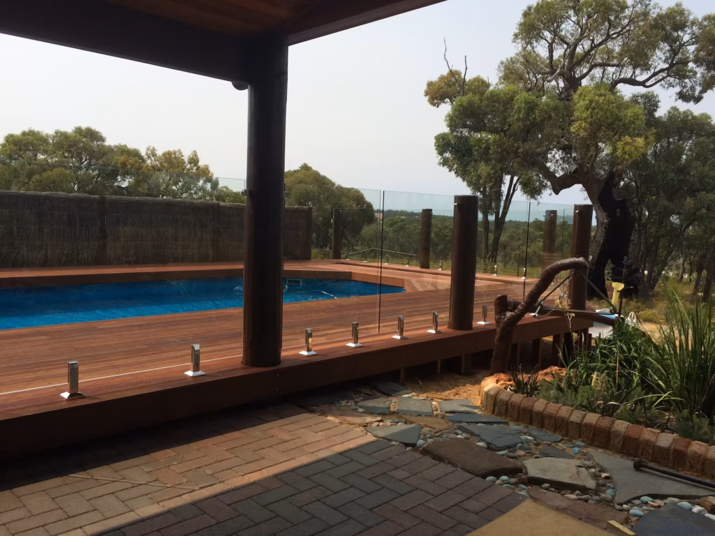 Frameless glass fence surrounding a wooden poolside area connected by wooden poles